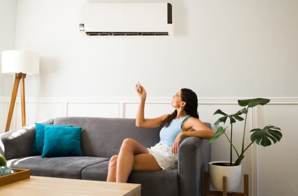 A woman sitting on a gray sofa points a remote control at a wall-mounted air conditioner in a living room. air conditioning service
