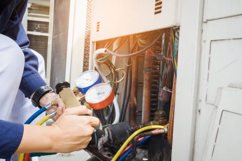 Technician uses pressure gauges to check and service an air conditioning unit, with wires and pipes visible inside the open panel.